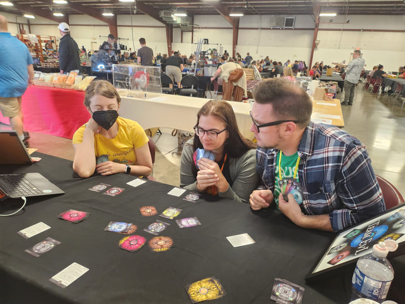 Three players leaning over a ritual circle at a convention table, the hall busy behind them