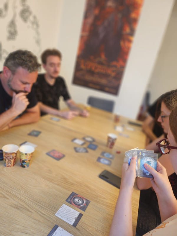 A game in progress around a long dining table, cards dealt out and a poster on the wall behind